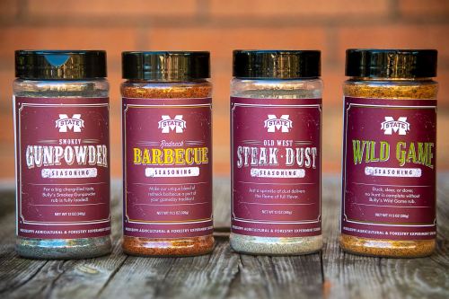 Four jars of seasoning: Smoky Gunpowder, Redneck Barbecue, Old West Steak Dust, and Wild Game, displayed on a wooden surface.