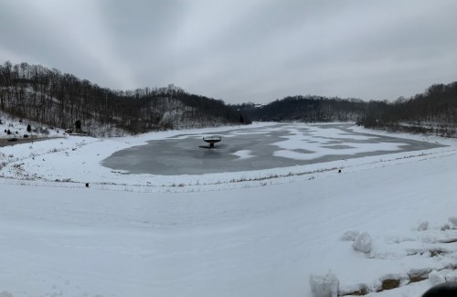 A snowy landscape with a frozen lake and a central stone structure, surrounded by wooded hills under a gray sky.