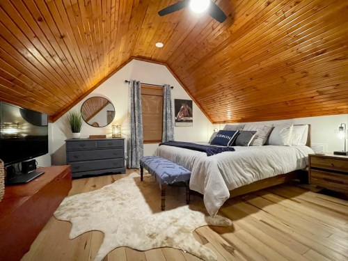 Cozy attic bedroom with wooden ceiling, a bed, dresser, and decorative rug, featuring warm lighting and stylish decor.
