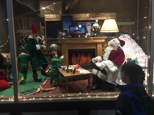 A festive scene with Santa, elves, and children in a cozy holiday setting, complete with a fireplace and decorations.