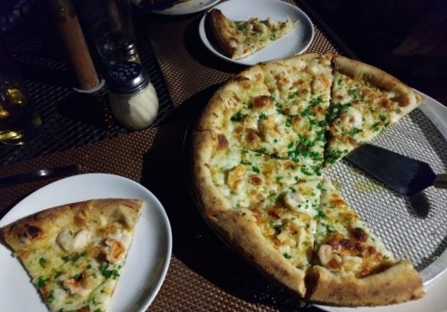 A large pizza topped with shrimp and herbs, with a slice on a plate beside it, set on a table with condiments.