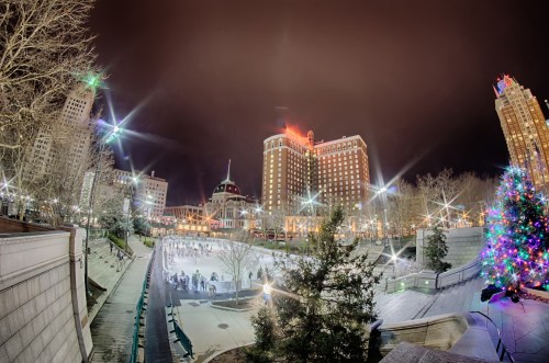 A festive ice skating rink surrounded by trees and buildings, illuminated by bright lights at night.