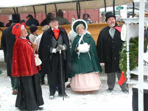 A group of four people in Victorian attire, enjoying a snowy outdoor event, with festive decorations around them.