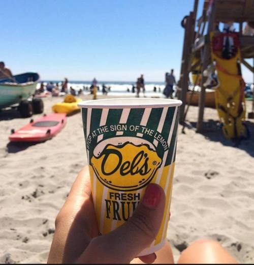 A hand holds a Del's Fresh Fruit lemonade cup on a sunny beach with people and boats in the background.