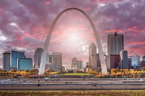 A sunset view of the Gateway Arch with a city skyline and colorful autumn trees in the foreground.