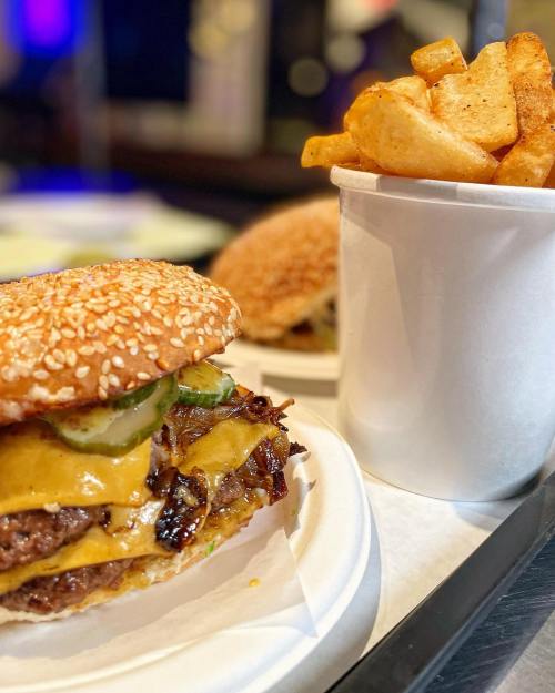 A close-up of a cheeseburger with pickles and crispy fries in a cup, served on a tray.