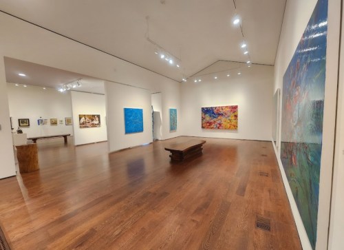 A spacious art gallery featuring colorful paintings on the walls and wooden flooring, with various artworks displayed.