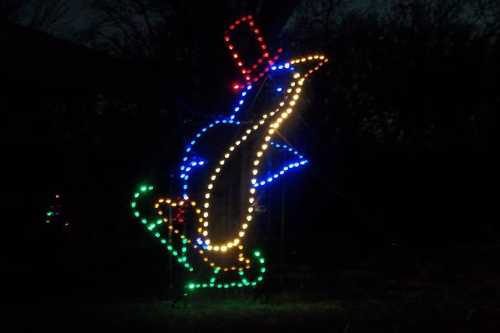 A lit-up penguin wearing a top hat and skating, surrounded by colorful lights against a dark background.