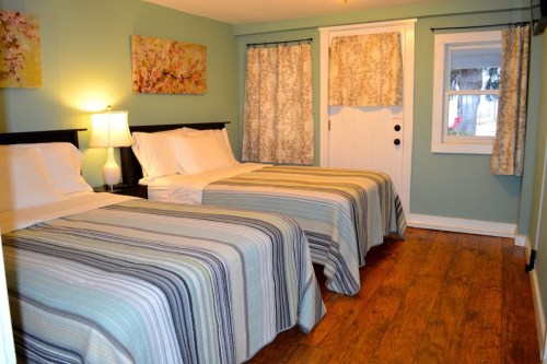 A cozy bedroom with two twin beds, light blue walls, floral artwork, and wooden flooring. Natural light from a window.