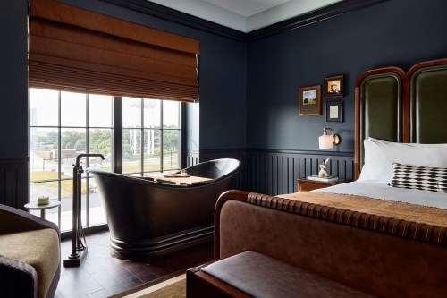A stylish hotel room featuring a large bathtub, a cozy bed, and a window with a view. Warm tones and modern decor.