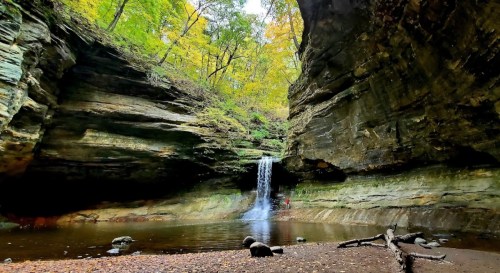 A serene waterfall cascades into a rocky pool, surrounded by lush green trees and steep canyon walls.