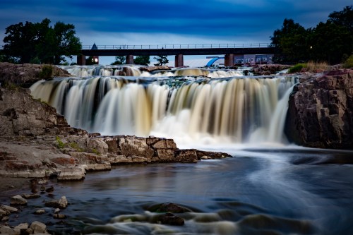 A serene waterfall cascades over rocky cliffs, with a bridge in the background under a twilight sky.
