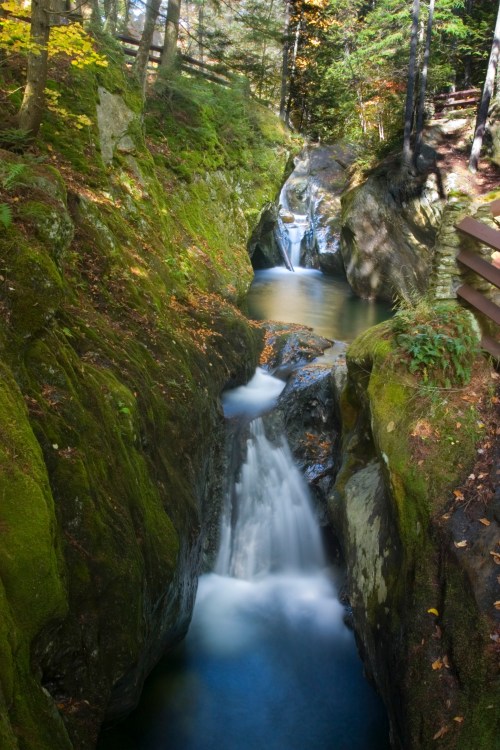 A serene waterfall cascades through a mossy, rocky gorge surrounded by trees in autumn colors.