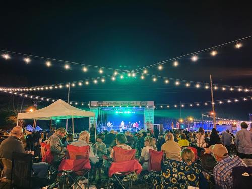 A lively outdoor concert at night, with a band on stage and an audience seated in folding chairs under string lights.