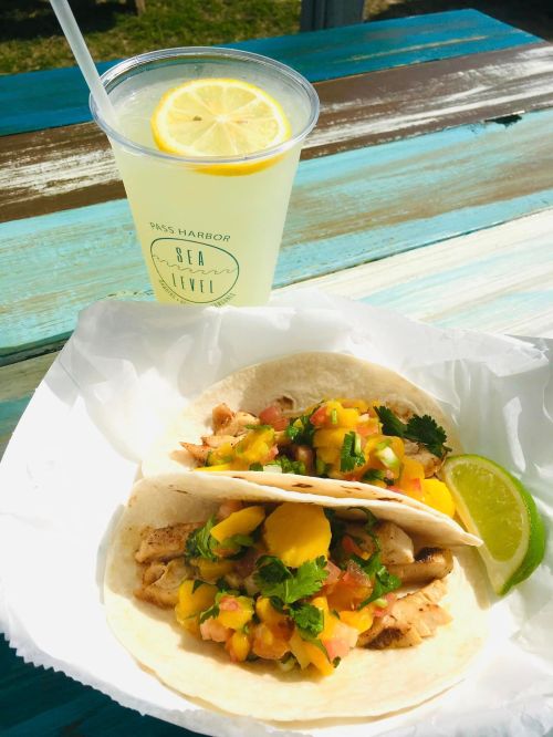 Two fish tacos topped with mango salsa and cilantro, served with a lime wedge, alongside a refreshing lemonade.
