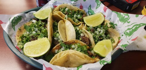 A platter of tacos topped with cilantro, onions, and lime wedges, served on a colorful paper liner.