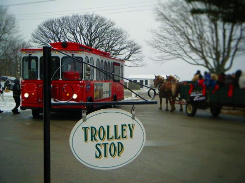 A red trolley at a stop sign, with a horse-drawn carriage in the background and bare trees nearby.
