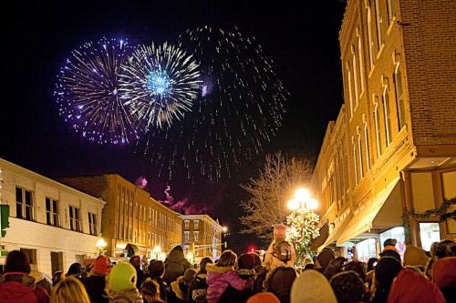 Crowd watching colorful fireworks display over a festive street at night, with buildings adorned in holiday decorations.