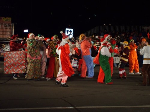 A festive group of musicians in colorful holiday costumes performing at night, surrounded by a cheering crowd.