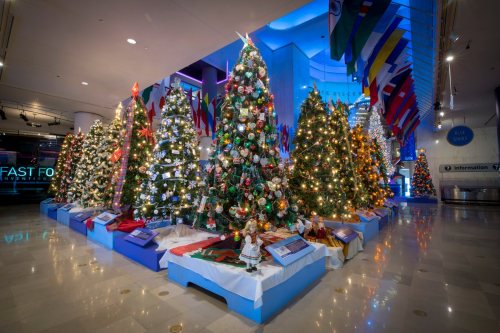 A festive display of decorated Christmas trees with lights and ornaments, surrounded by flags in a modern indoor space.