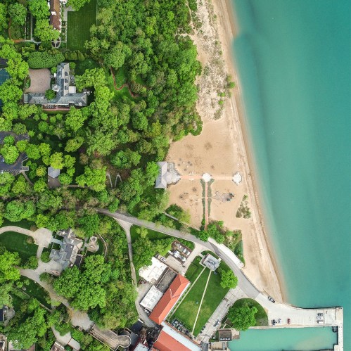 Aerial view of a lush green area with a sandy beach and blue water, featuring homes and pathways nearby.