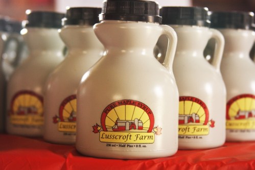 Bottles of Lusscroft Farm pure maple syrup on a red tablecloth, featuring a sun logo and farm illustration.