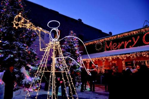 A glowing angel figure with a trumpet stands among decorated trees in a festive outdoor setting at dusk.