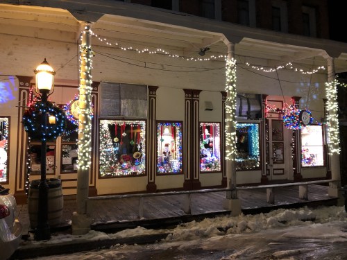 Festively decorated storefronts with colorful lights and wreaths, surrounded by snow on a winter evening.