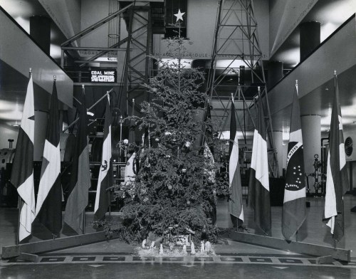 A Christmas tree surrounded by various flags in a large indoor space, decorated for the holiday season.