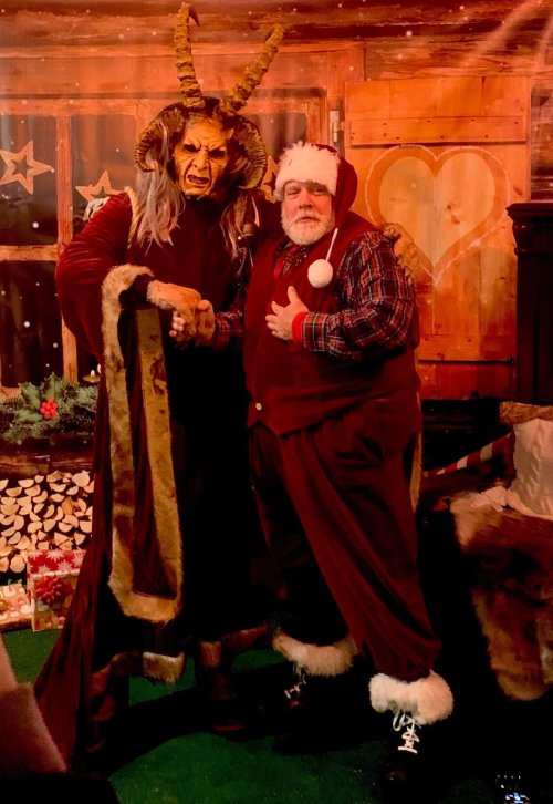 A man in a Santa costume poses with a figure in a horned, demonic costume in a festive setting.