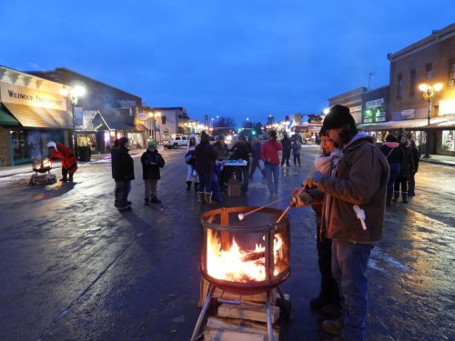 People gather around a fire pit on a snowy street, enjoying the evening atmosphere and roasting marshmallows.