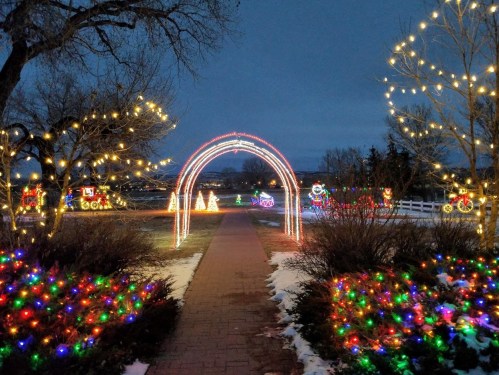 A festive pathway adorned with colorful lights, leading to decorated trees and snowmen in a winter landscape.