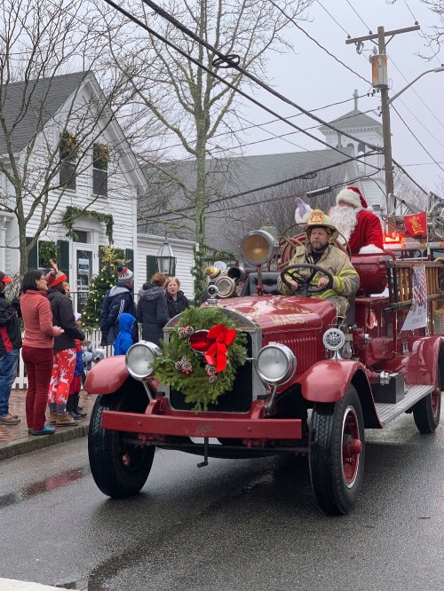 A vintage fire truck decorated for Christmas, with Santa and a firefighter driving, surrounded by festive onlookers.