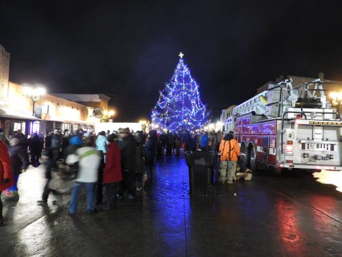 A festive crowd gathers around a large, illuminated Christmas tree, with a fire truck parked nearby on a dark, rainy night.