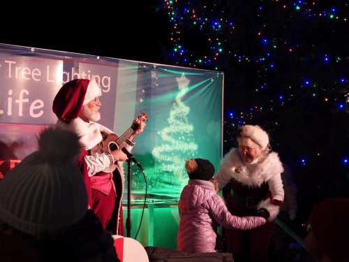 A festive scene with Santa playing guitar, a child dancing, and a woman in a holiday outfit near a decorated tree.