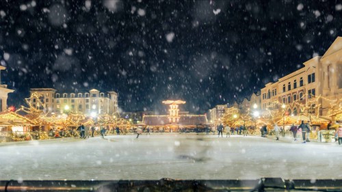 A snowy night scene of a festive plaza, adorned with lights and decorations, bustling with people enjoying the holiday atmosphere.