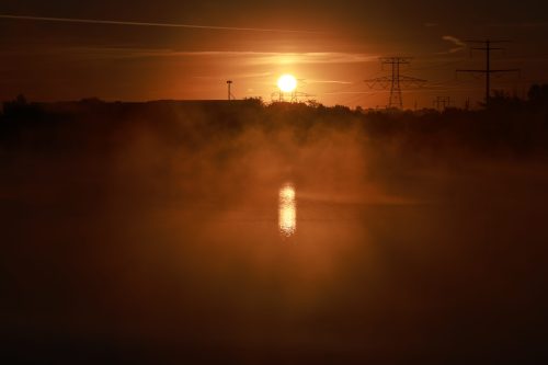 Sunset over a misty lake, with silhouettes of power lines and trees against a vibrant orange sky.