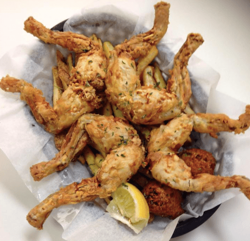 A platter of fried frog legs served with fries, a lemon wedge, and a side of hushpuppies on parchment paper.