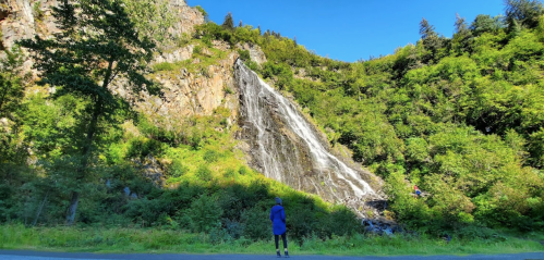 A person stands in front of a tall waterfall cascading down a rocky cliff, surrounded by lush greenery and blue sky.