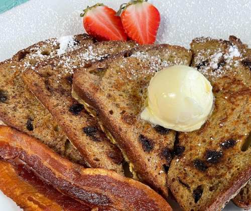 Plate of French toast topped with butter, powdered sugar, and served with crispy bacon and fresh strawberries.