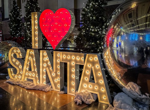 Brightly lit sign reading "I ❤️ SANTA" surrounded by festive decorations and large reflective ornaments.