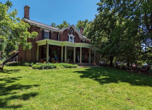 A large brick house with a porch, surrounded by green grass and trees on a sunny day.