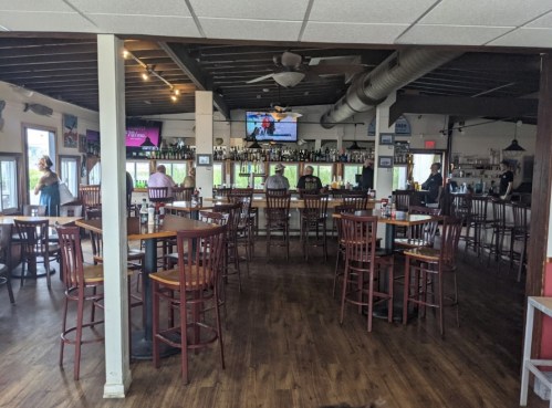 Interior of a restaurant with wooden tables and chairs, a bar area, and a TV showing a sports event.