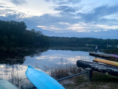 A serene lake at dusk, with kayaks lined up and a person fishing in the distance under a cloudy sky.