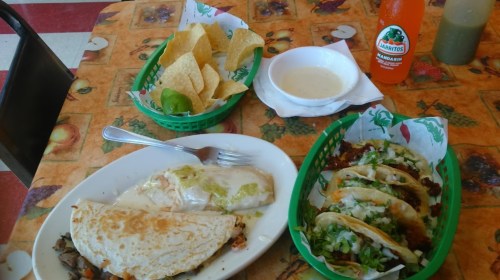 A colorful table with tacos, a quesadilla, chips with salsa, and a bottle of soda.