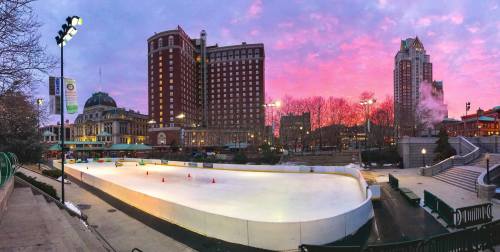 A panoramic view of an ice skating rink at sunset, surrounded by buildings and trees, with vibrant pink and purple skies.