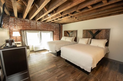 Cozy hotel room with two beds, exposed brick walls, wooden beams, and large windows letting in natural light.