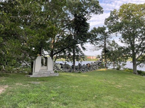 A serene park scene featuring a stone monument near a river, surrounded by trees and grassy areas.