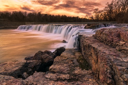 A serene waterfall cascades over rocks at sunset, with vibrant colors reflecting in the water and trees in the background.