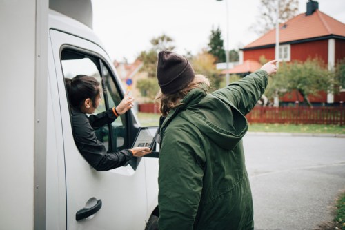 A person in a van points while another person outside gestures, both looking towards a nearby house.